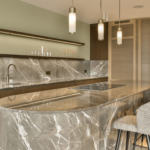 A modern kitchen with marble counter tops and bar stools in the center of the room is light green walls