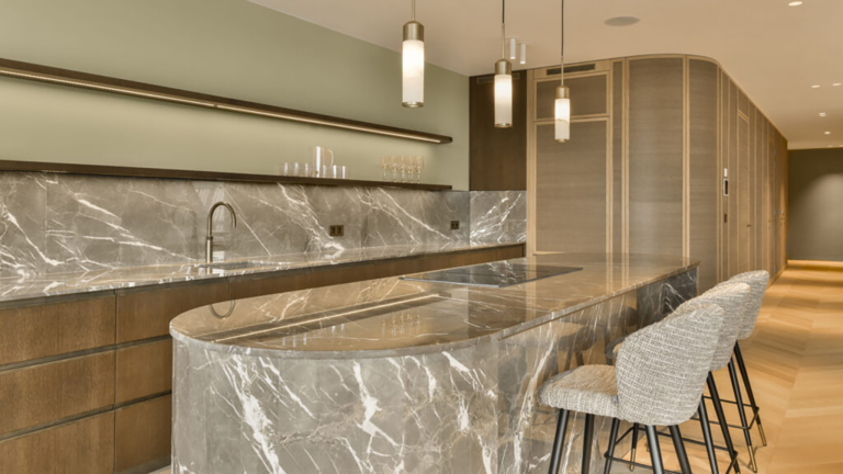 A modern kitchen with marble counter tops and bar stools in the center of the room is light green walls