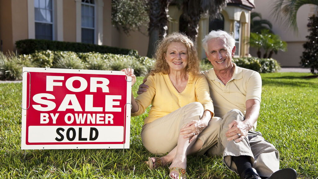 Senior couple in front of a house with a For Sale – Sold sign – mortgages for retirees