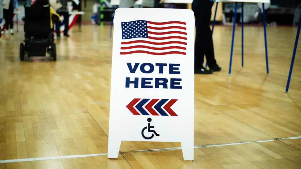 Tabliczka Vote Here z amerykańską flagą w lokalu wyborczym – wybory 3 listopada 2026