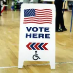 Tabliczka Vote Here z amerykańską flagą w lokalu wyborczym – wybory 3 listopada 2026